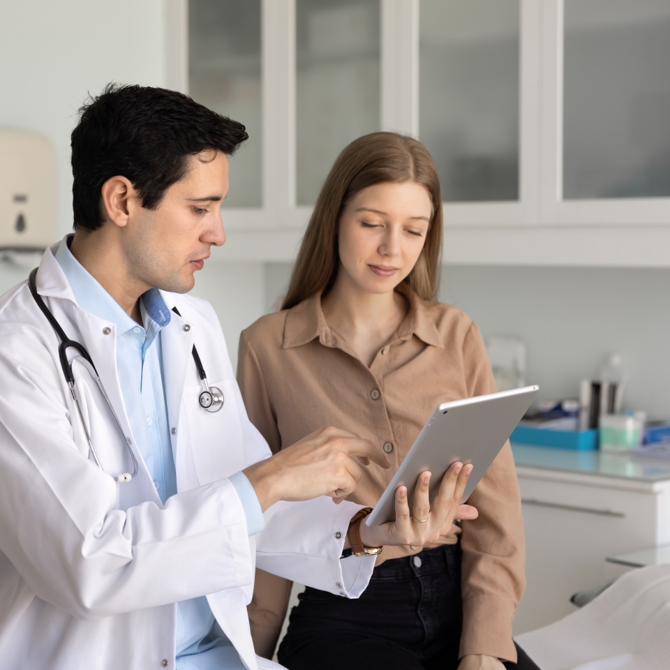 Male physician explains medical information to young female patient using digital tablet in clinic, showing CT scan imaging results, sharing treatment plan, discussing new telemedicine platform or app