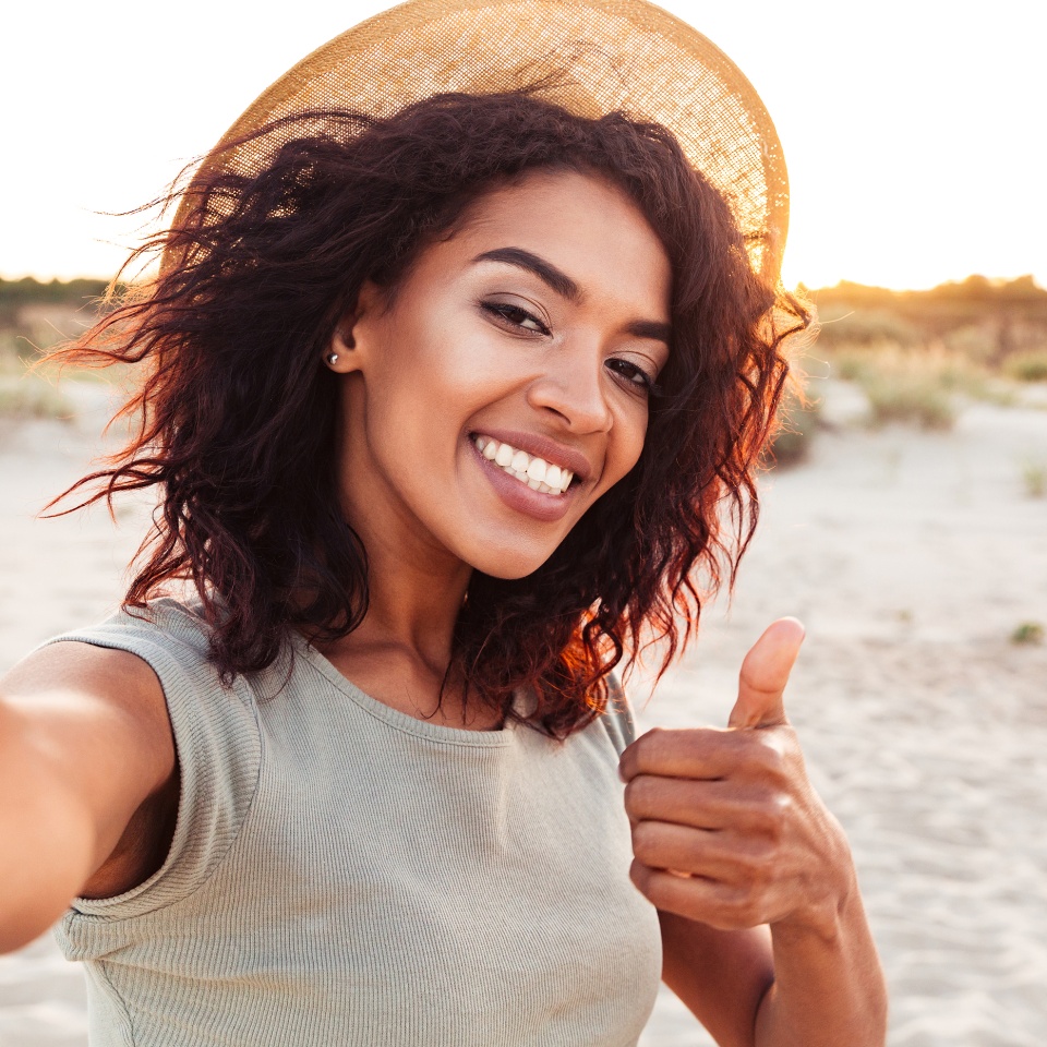 Close up of cheerful young african girl in summer hat taking a selfie at the beach and showing thumbs up gesture