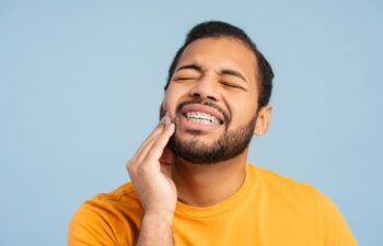 Portrait of troubled, african american man with braces, holding his cheek in pain, against a blue background. Illustrates dental discomfort concept
